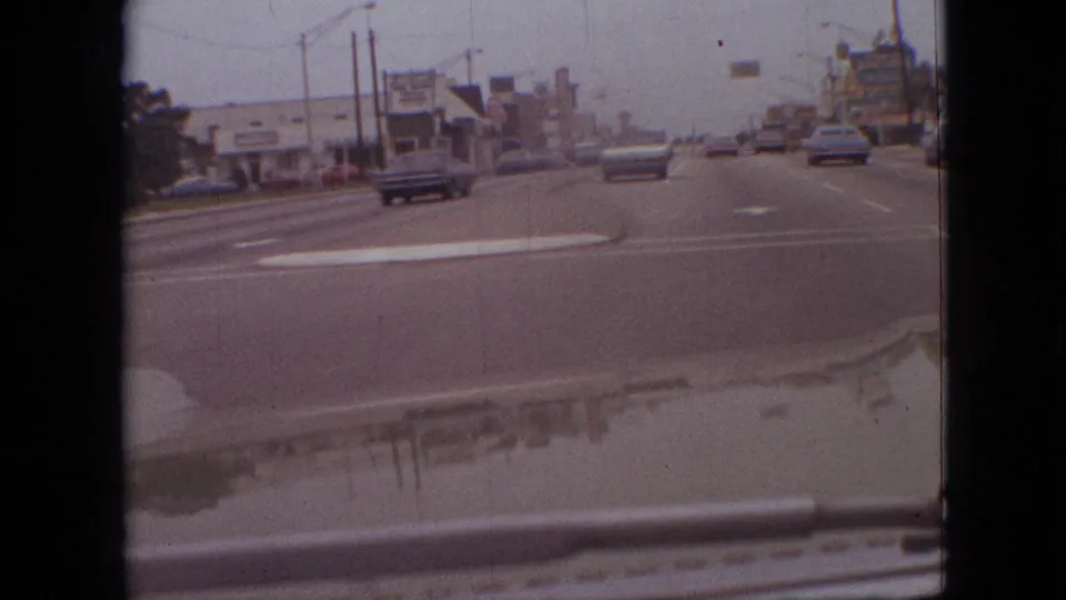 First-person driving view in Branson, Missouri, showing cars ahead on a broad 1968 roadway, low commercial buildings, overhead streetlights, and visible 8mm gate edges.