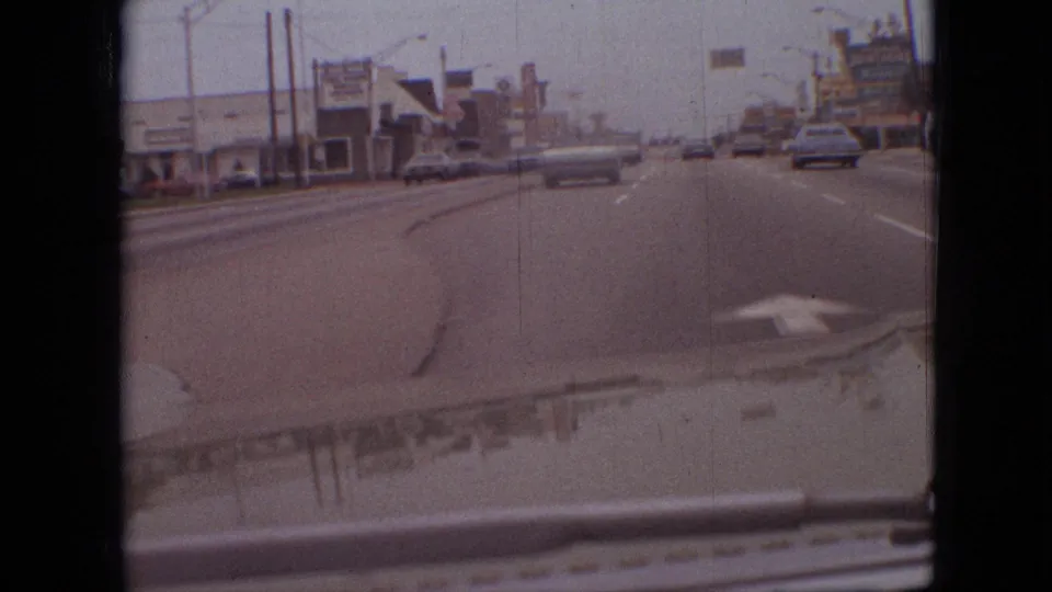 1968 Branson, Missouri home-movie frame showing a forward view over a car hood along a multi-lane commercial road with cars and roadside signs.