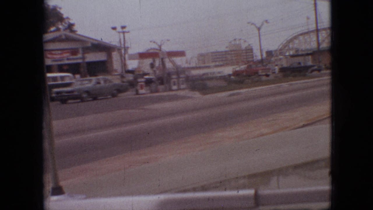 1968 Branson, Missouri frame showing a service station beside a multi-lane road, with cars in traffic and an amusement-park roller coaster beyond.