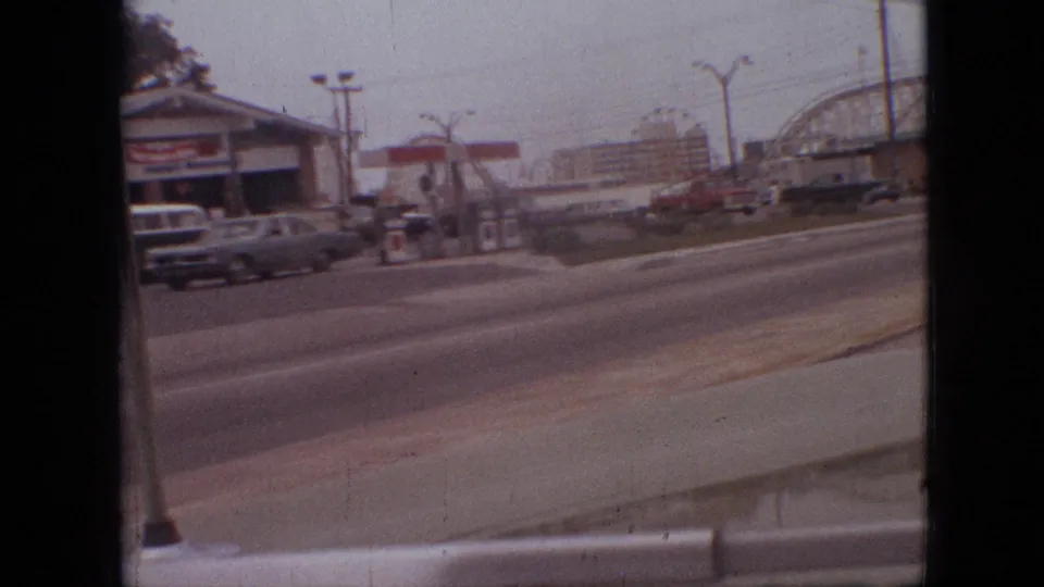 1968 Branson, Missouri frame showing a service station beside a multi-lane road, with cars in traffic and an amusement-park roller coaster beyond.