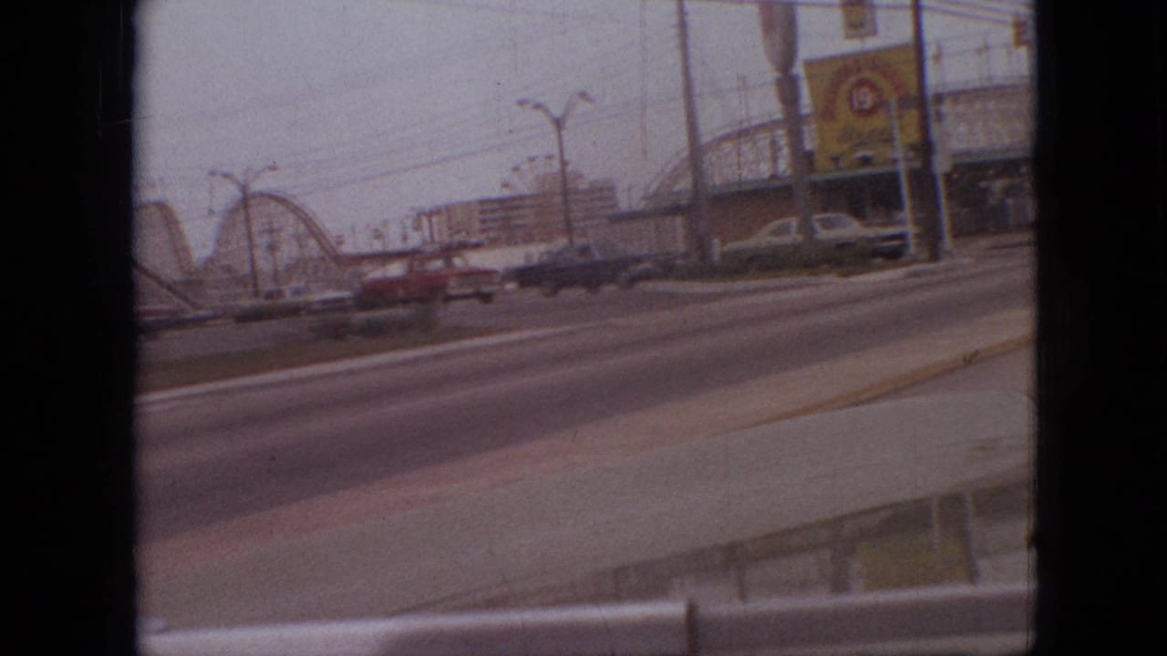 1968 Branson, Missouri home-movie frame showing a roller coaster and attraction signage across a multi-lane road with cars passing in the foreground.