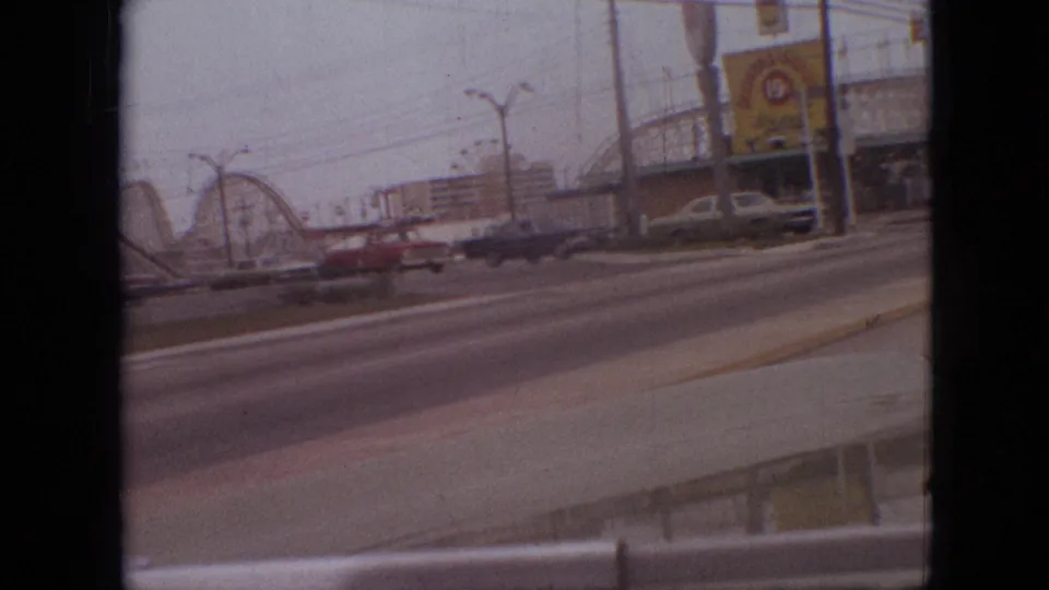 1968 Branson, Missouri home-movie frame showing a roller coaster and attraction signage across a multi-lane road with cars passing in the foreground.