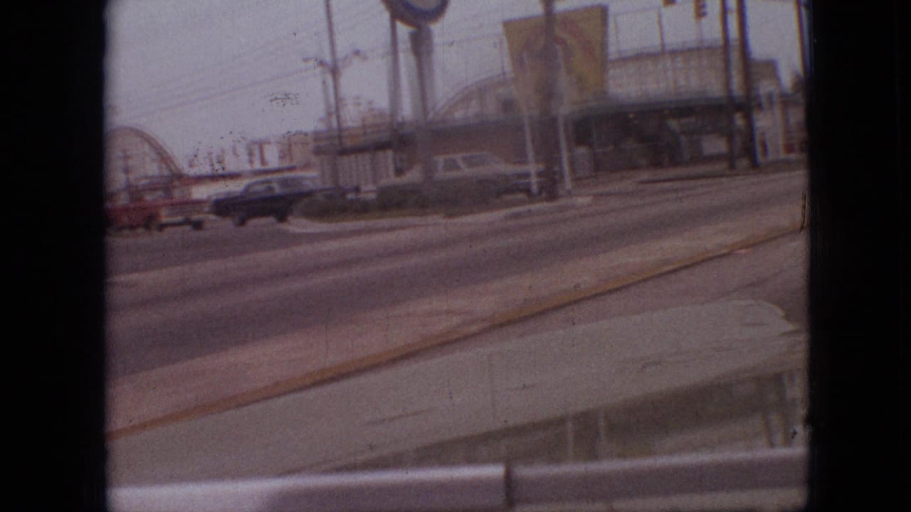 1968 Branson, Missouri home-movie frame showing a roadside intersection from inside a vehicle, with passing cars, signs, and window or film-frame edges visible.