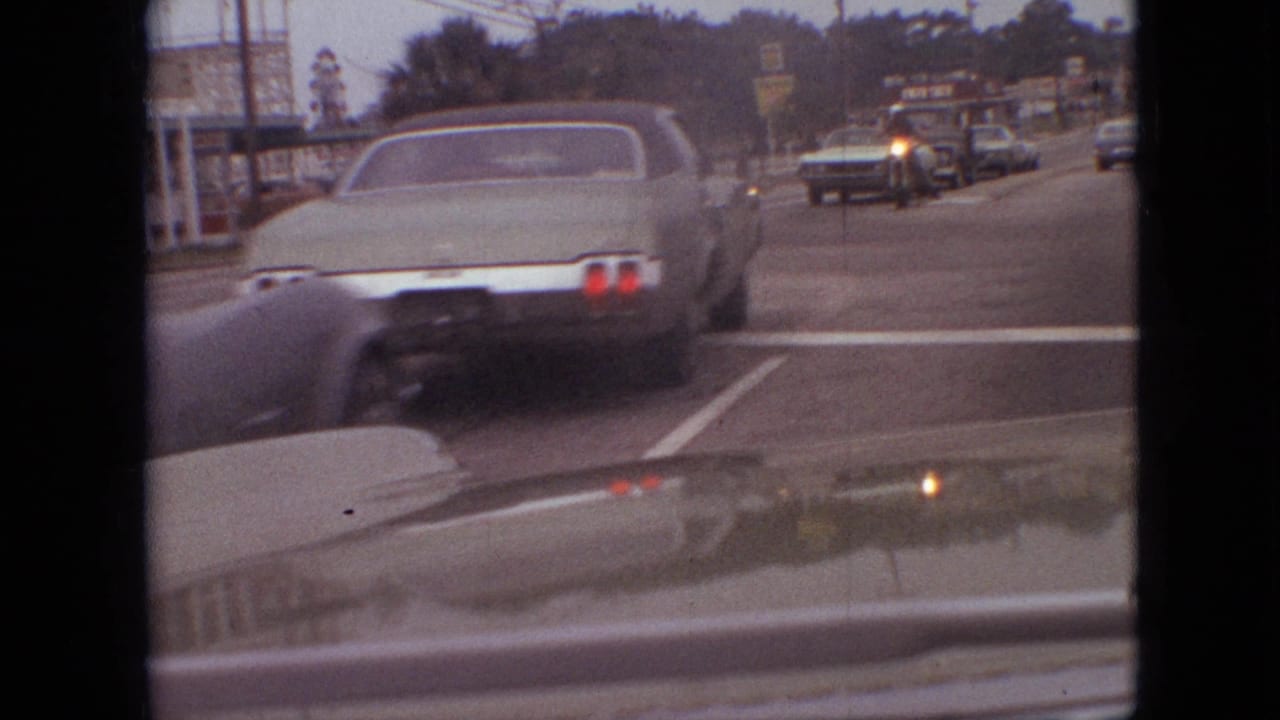 Green sedan ahead at an intersection with oncoming traffic and roadside signs in the distance.