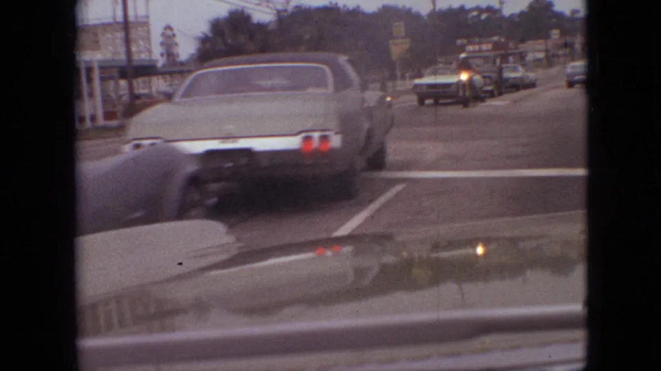 1968 Branson, Missouri traffic view from a moving vehicle, with a green sedan in front, oncoming cars, road markings, and distant commercial signage.