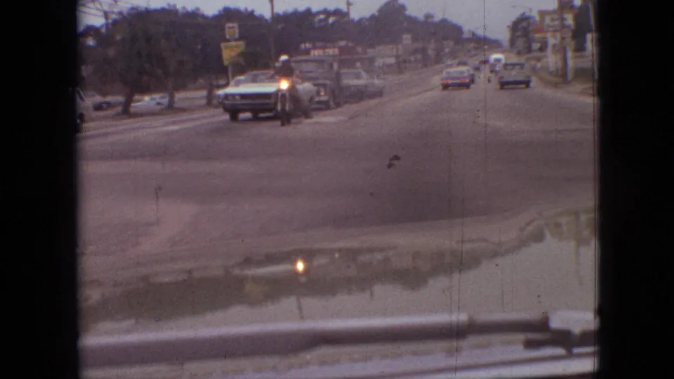 1968 Branson, Missouri street traffic seen from inside a car, including approaching vehicles, a motorcycle or small motorbike, roadside signs, and soft 8mm motion blur.