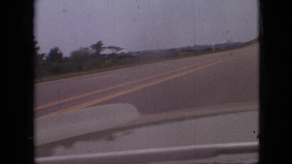 1968 Branson, Missouri driving frame showing an open two-lane road from a moving vehicle, with double yellow lines, roadside vegetation, and distant low hills.