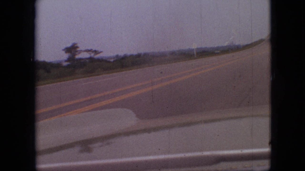 1968 Branson, Missouri home-movie frame showing a curving two-lane road from inside a moving car, with double yellow centerlines, roadside vegetation, and an overcast sky.