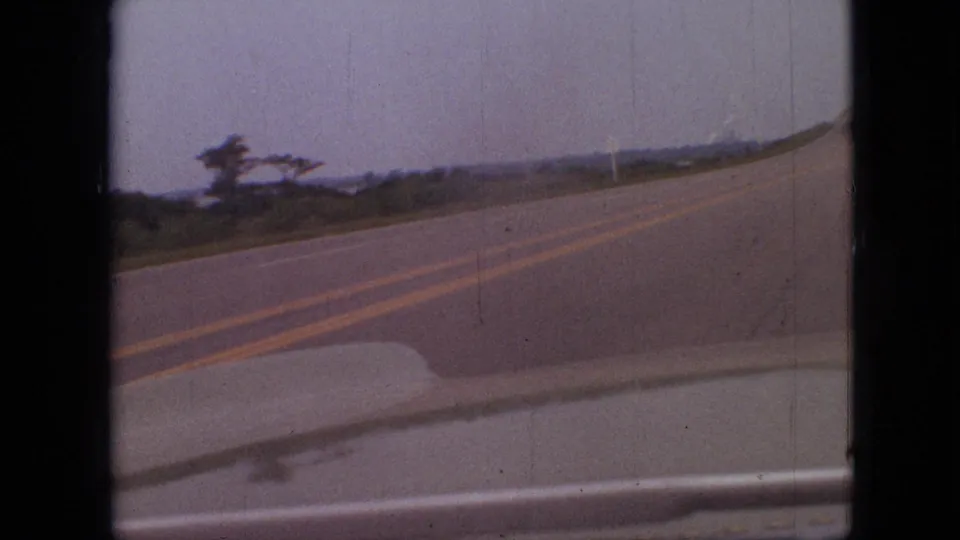 1968 Branson, Missouri home-movie frame showing a curving two-lane road from inside a moving car, with double yellow centerlines, roadside vegetation, and an overcast sky.