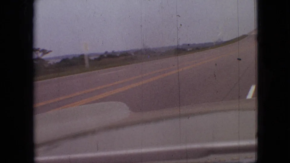 1968 Branson, Missouri road-trip frame from a moving vehicle, showing a paved two-lane road bending ahead through low roadside terrain under muted sky.