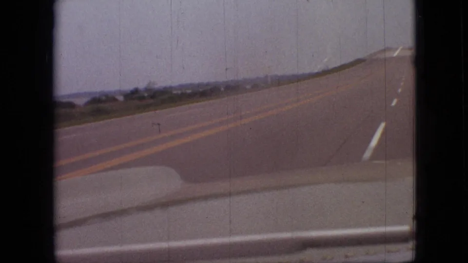 1968 Branson, Missouri home-movie still showing a passenger-side view of a two-lane roadway, with yellow center markings, a white shoulder line, and distant roadside trees.