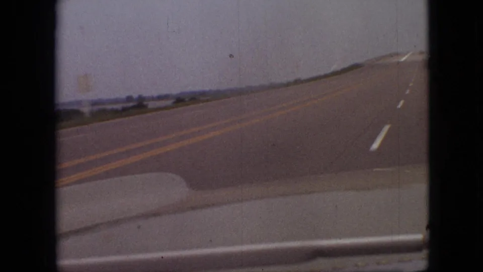 1968 Branson, Missouri travel frame showing a rural two-lane road curving away from the car, with double yellow lines, roadside greenery, and soft 8mm film wear.