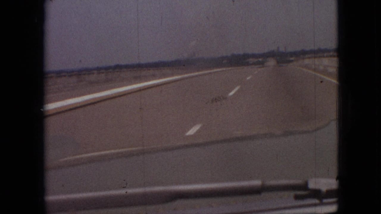 1968 8mm home-movie frame from near Branson, Missouri, showing a forward car view along a wide flat highway with lane markings and a low distant horizon.