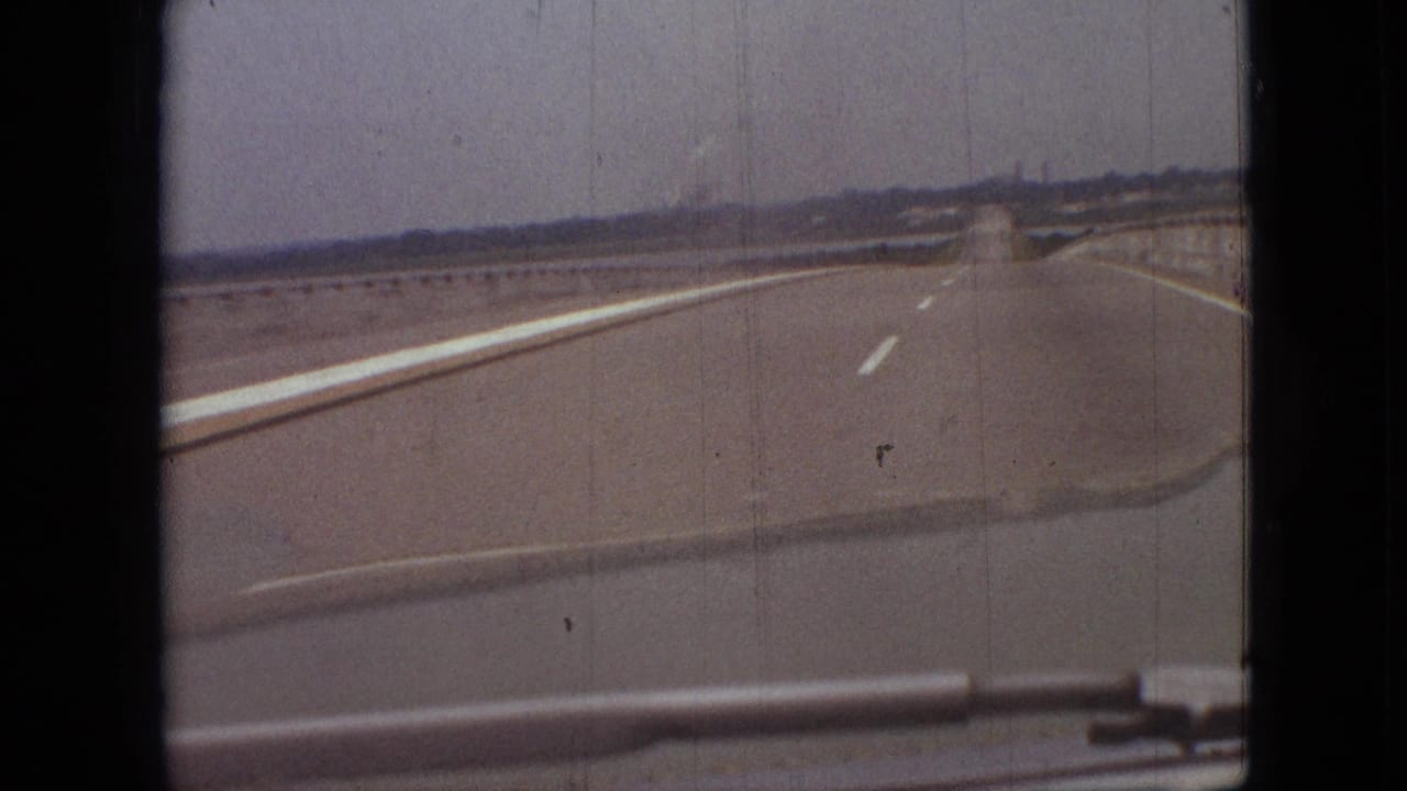 1968 8mm frame from the Branson, Missouri road scene, showing the highway continuing ahead with a faint smoke plume and distant roadside forms on the horizon.