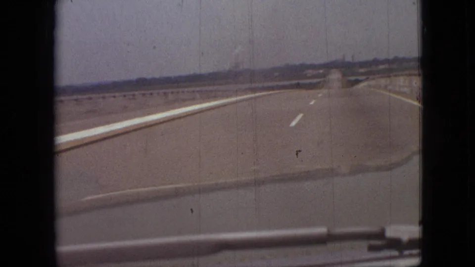 1968 8mm frame from the Branson, Missouri road scene, showing the highway continuing ahead with a faint smoke plume and distant roadside forms on the horizon.