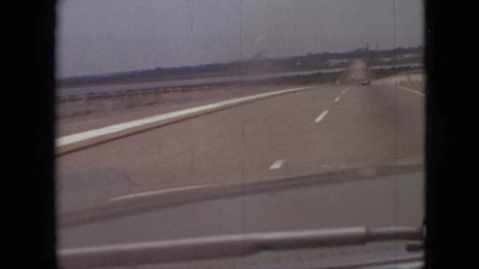 1968 8mm home-movie frame showing a forward road view near Branson, Missouri, with flat pavement, painted lane lines, low horizon structures, and soft film grain.
