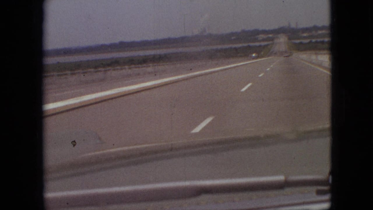 1968 Branson, Missouri home-movie view from inside a moving car looking down a wide paved highway with white lane markings, low barriers, and distant open terrain.