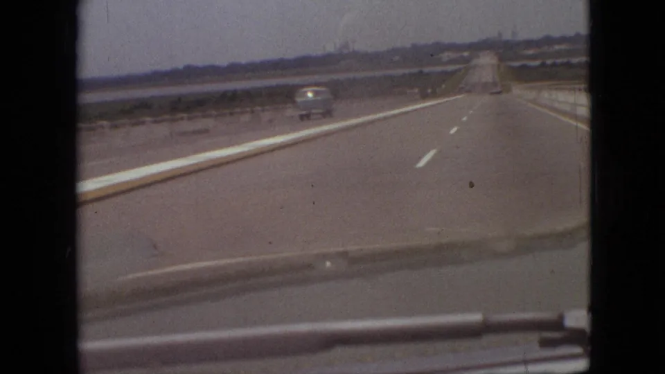 1968 Branson, Missouri car-window view on a multi-lane road with a light-colored vehicle ahead, roadside barriers, open terrain, and distant trees.
