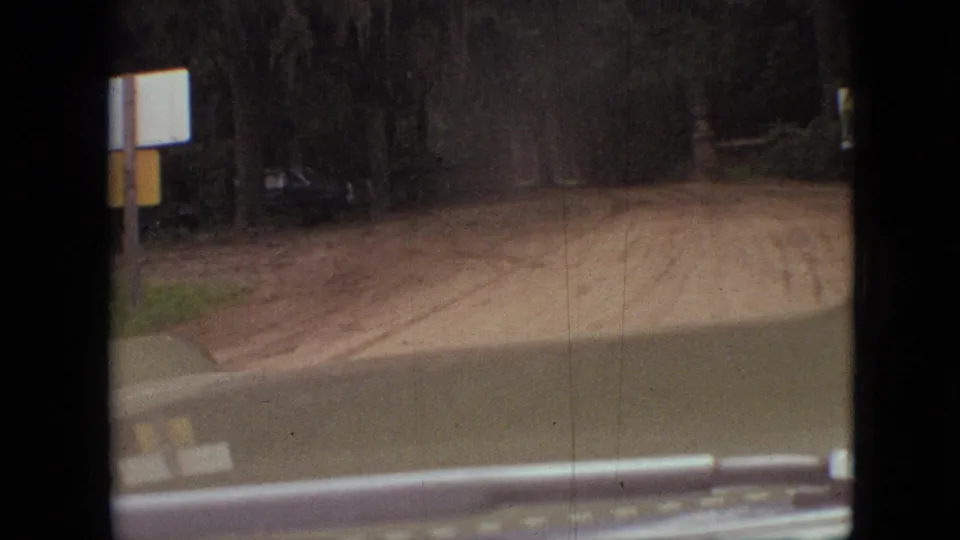 1968 Branson, Missouri home-movie frame showing a turn or approach toward a dirt road bordered by dense trees, with a roadside sign shape and vehicle windshield edge in view.