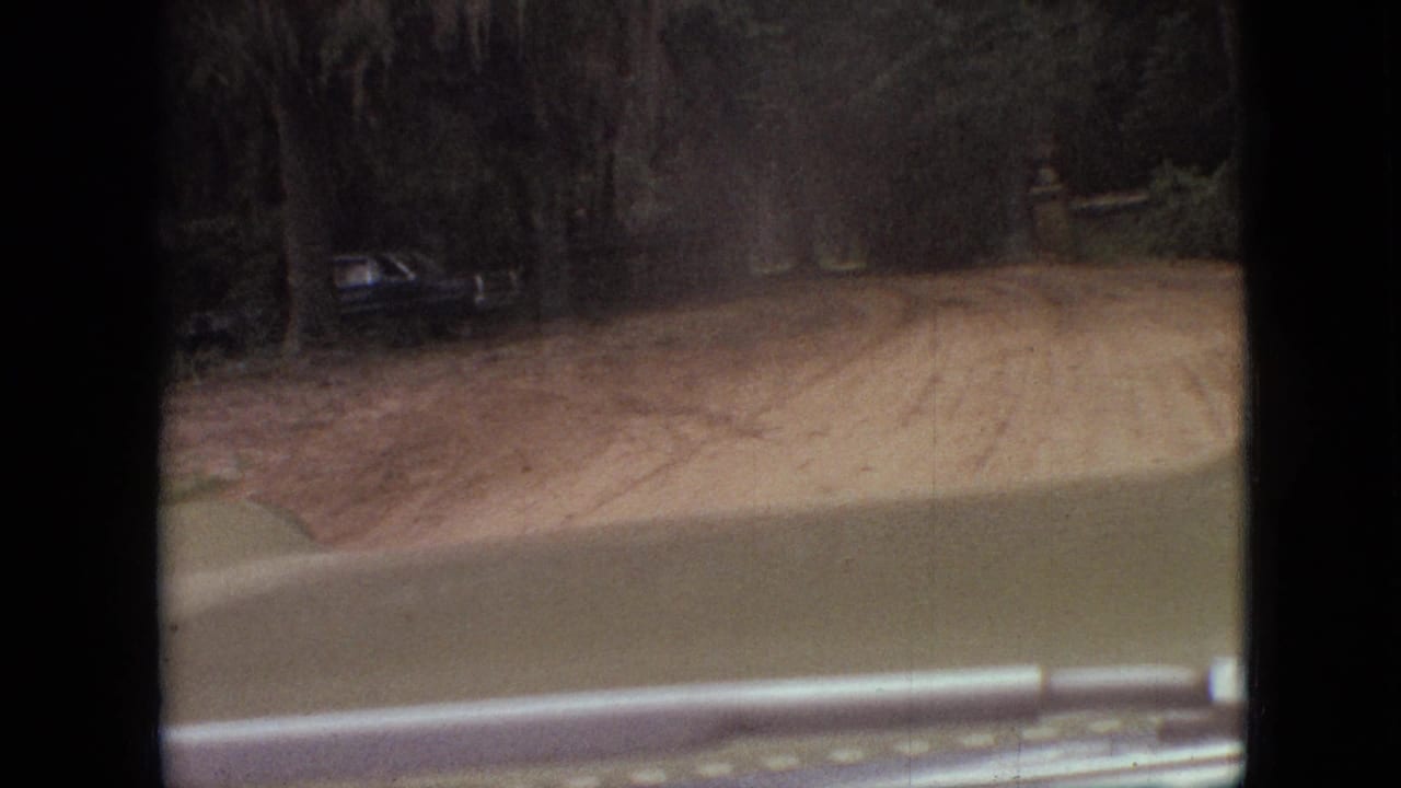 1968 Branson, Missouri 8mm frame showing a passenger-side windshield view over the car hood toward a winding dirt road bordered by trees, with a dark vehicle partly visible on the left.
