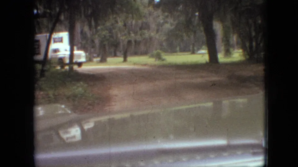 1968 Branson, Missouri 8mm frame looking over the car hood toward a wooded dirt road, with a white truck or van and roadside signs visible on the left.