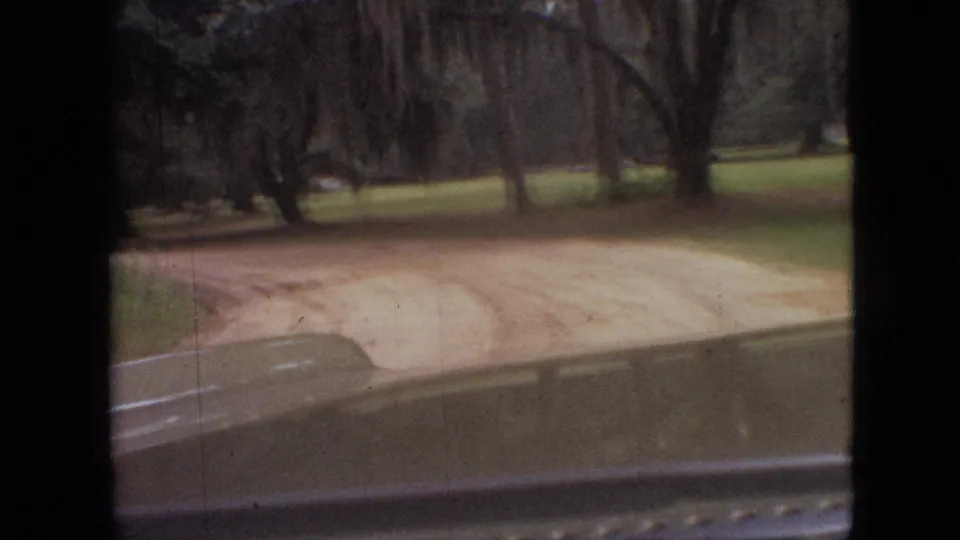 1968 Branson, Missouri home-movie frame looking from a moving car toward a curving dirt road bordered by grass and moss-draped trees.