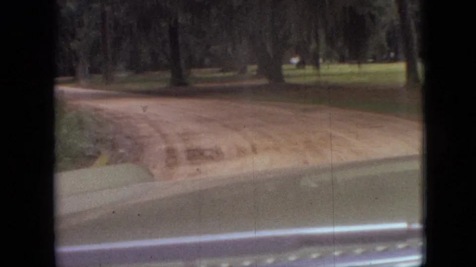 1968 Branson, Missouri home-movie frame showing a dirt road crossing the view from inside a car, with trees and grass beyond the windshield.