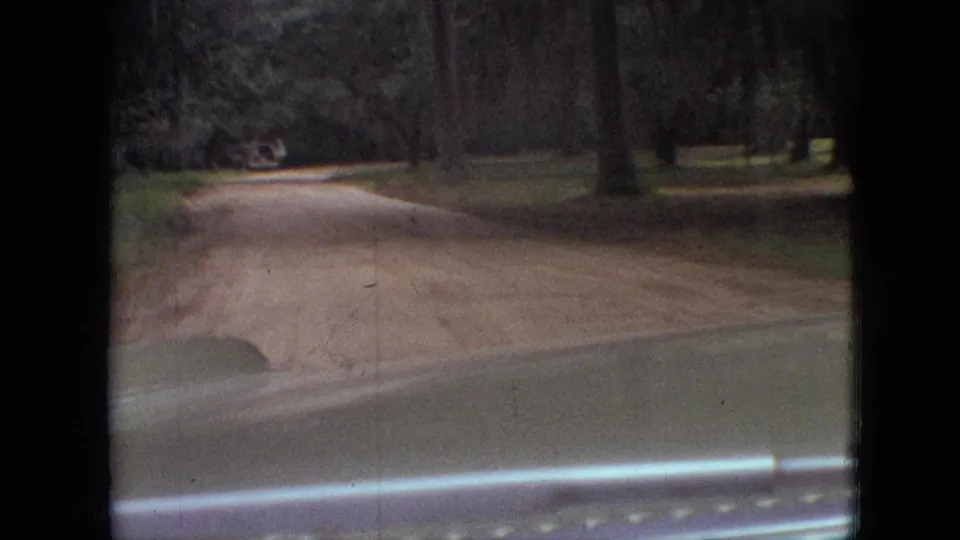 1968 Branson, Missouri home-movie frame looking down a dirt road through trees from inside a car, with the hood and windshield edge in the foreground.