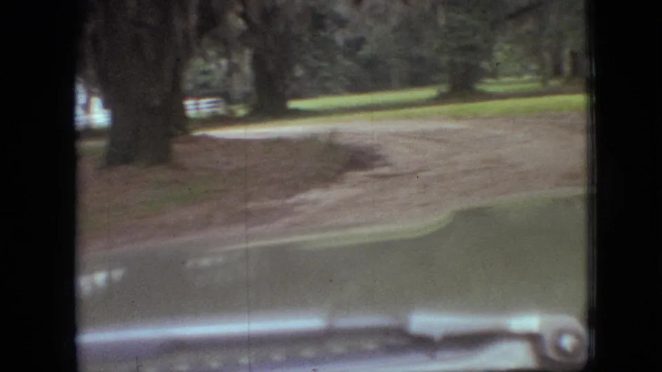 The car passes a curving dirt road or driveway beneath large trees, with grass and the windshield edge visible in the 1968 Branson footage.