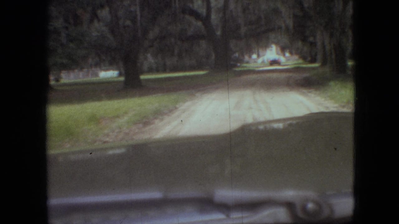 1968 Branson, Missouri home-movie frame showing a car windshield view toward a pale dirt road bordered by grass and large trees.