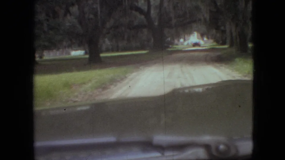 1968 Branson, Missouri home-movie frame showing a car windshield view toward a pale dirt road bordered by grass and large trees.
