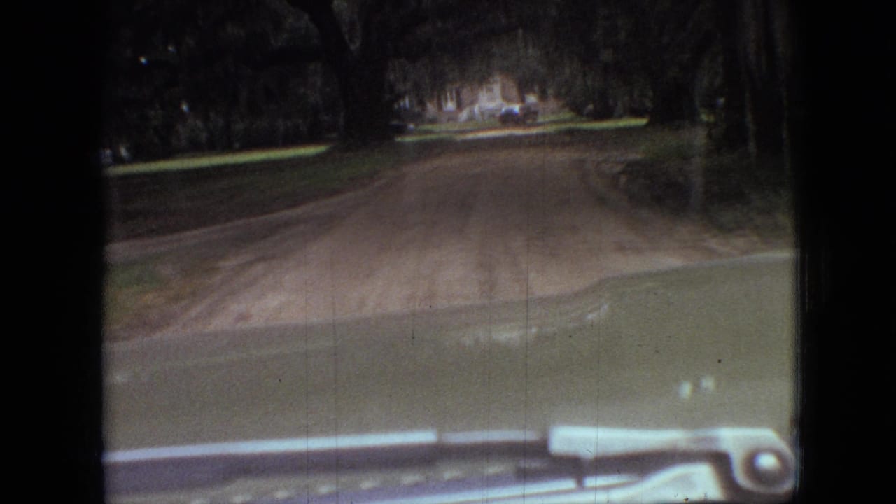Dirt road seen through a car windshield with trees and a house ahead.