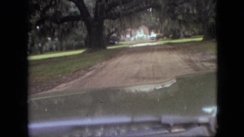 1968 Branson, Missouri home-movie still showing the car nearing a wider stretch of dirt road with lawn, trees, and a house in the distance.