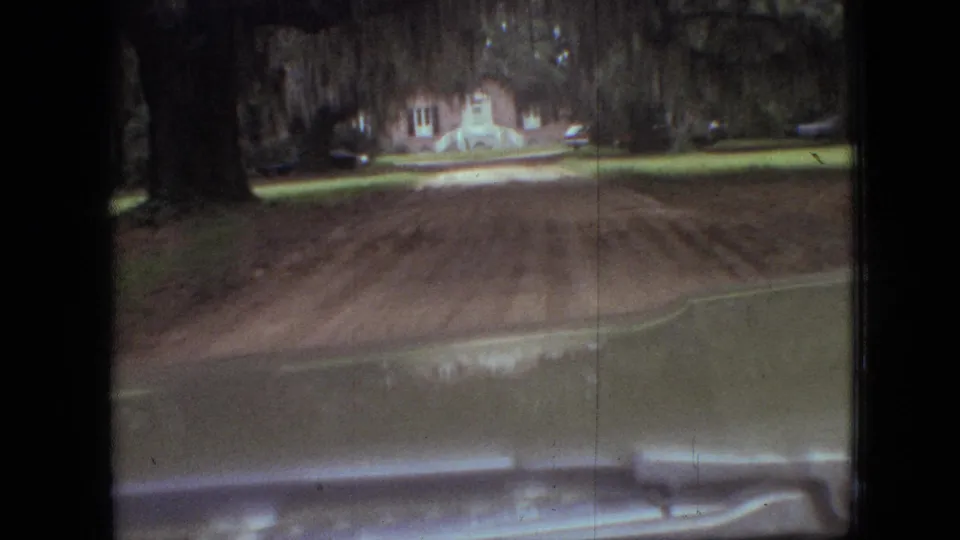 1968 Branson, Missouri frame showing a dashboard-level car POV across an unpaved road toward grass, mature trees, and a distant house.