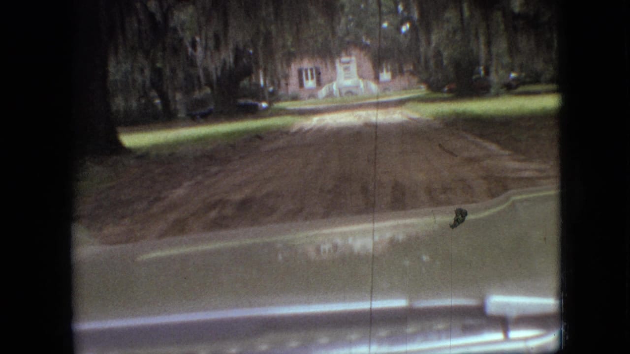 1968 Branson, Missouri 8mm frame showing a vehicle windshield view toward a brick house, dirt driveway, lawn, parked car, and large trees with hanging moss.