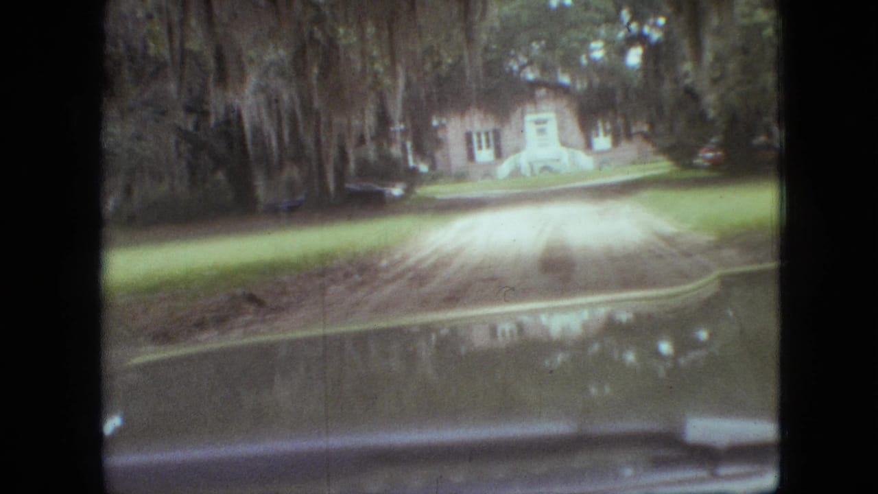 Vehicle-view 8mm frame from Branson, Missouri, 1968, with a dirt driveway, grass, brick house, white steps, trees, and a parked car visible through the windshield.