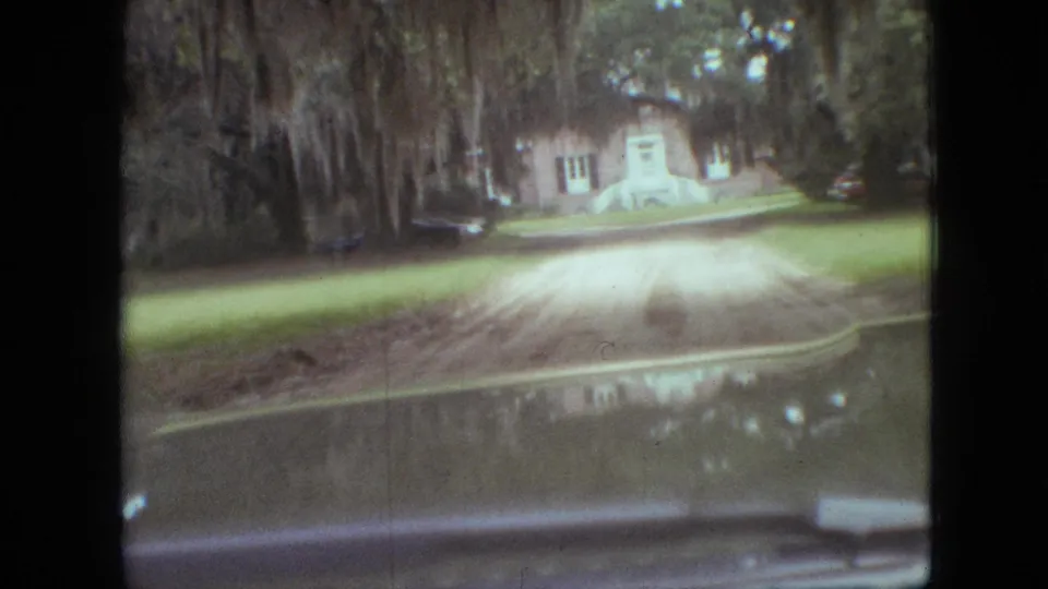 Vehicle-view 8mm frame from Branson, Missouri, 1968, with a dirt driveway, grass, brick house, white steps, trees, and a parked car visible through the windshield.