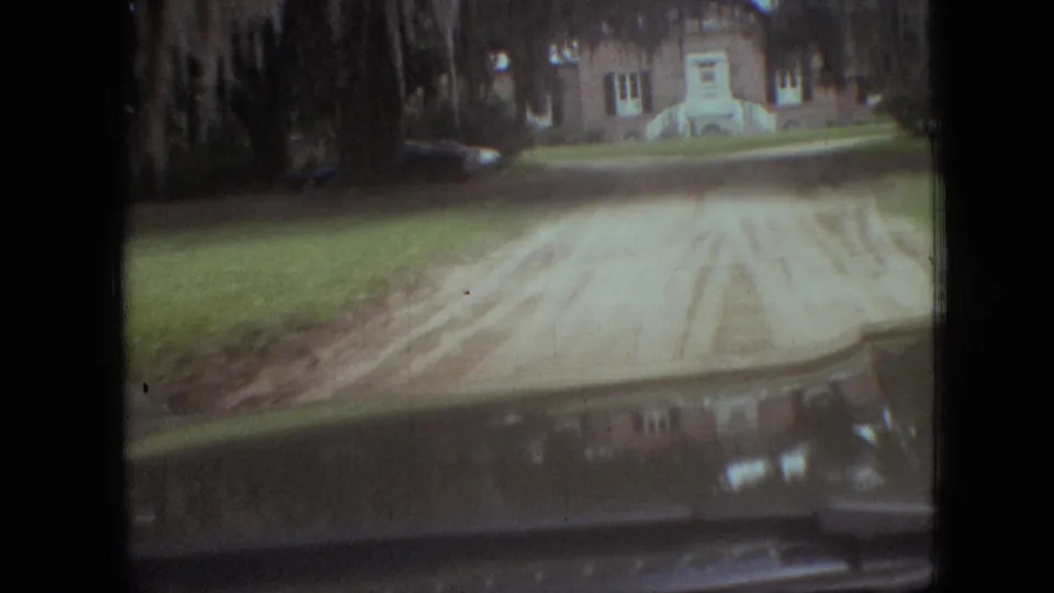 Closer 1968 Branson, Missouri home-movie frame showing the driveway and lawn in front of a brick house, with trees and parked cars partly visible through the windshield.