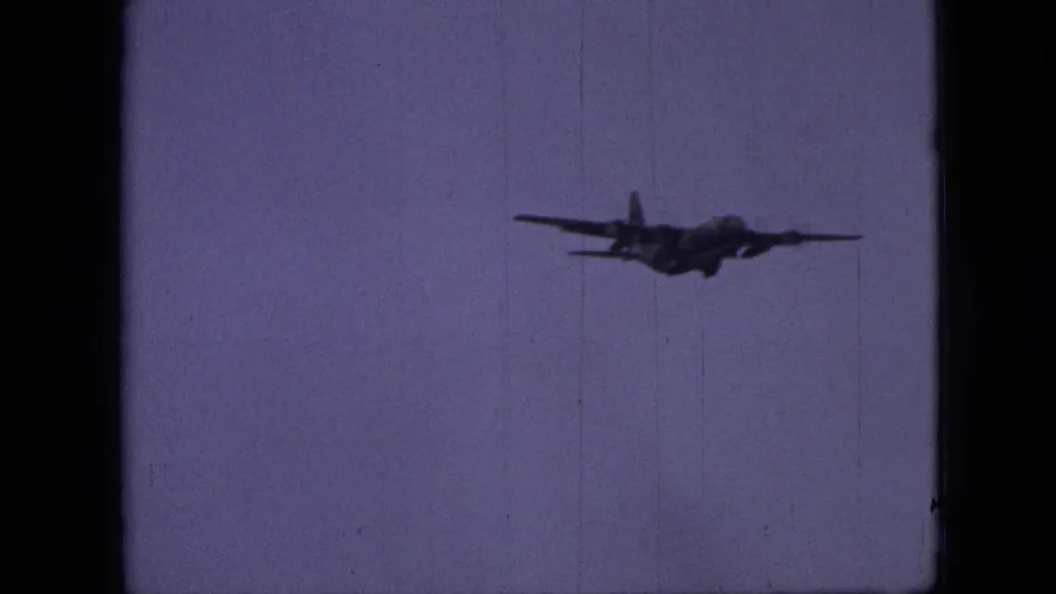 1968 8mm frame showing a dark airplane silhouette flying across a purple-tinted sky, with film grain and vertical scratches visible.