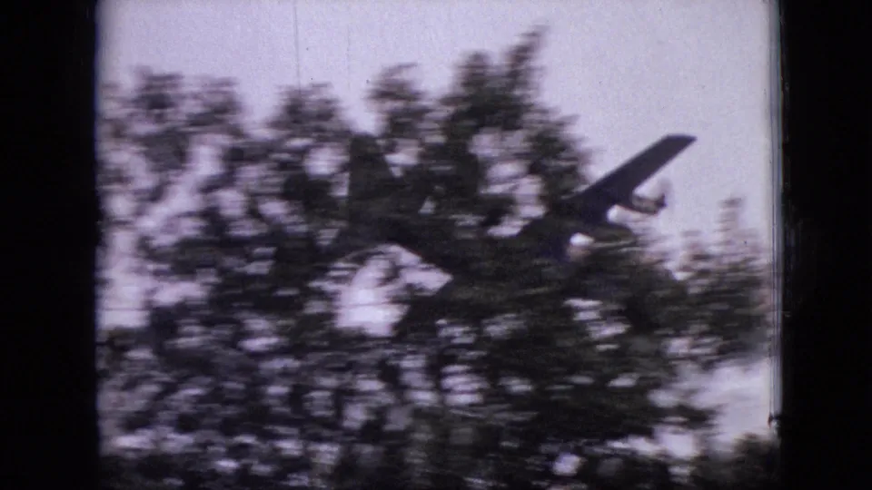 A small propeller airplane emerges above the treeline in metadata-supported 1968 Branson, Missouri home-movie footage, framed by soft grain and dark scan edges.