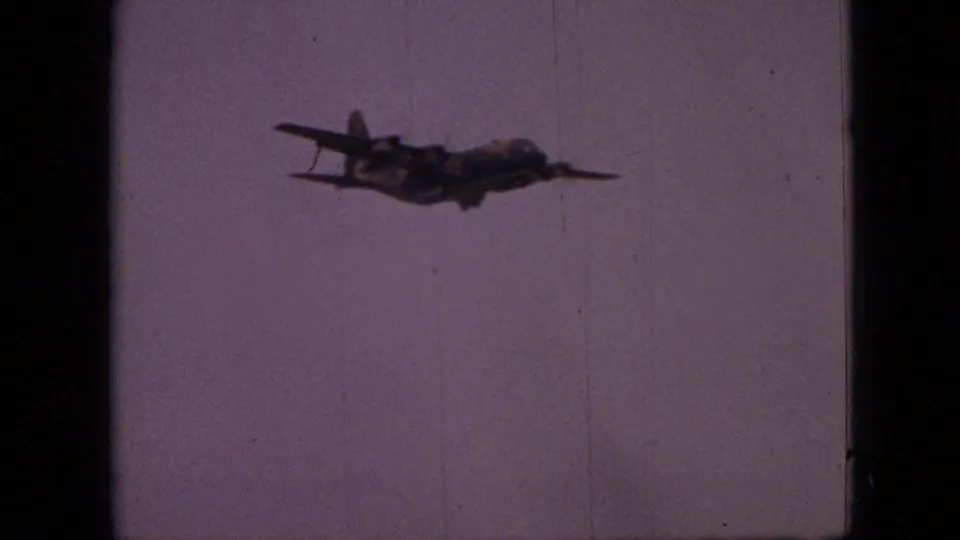 A military-style transport aircraft flies across a pale purple sky in 1968 Branson, Missouri home-movie footage.