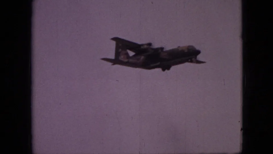 The transport aircraft appears in side view against a pale sky, framed by dark 8mm film edges.