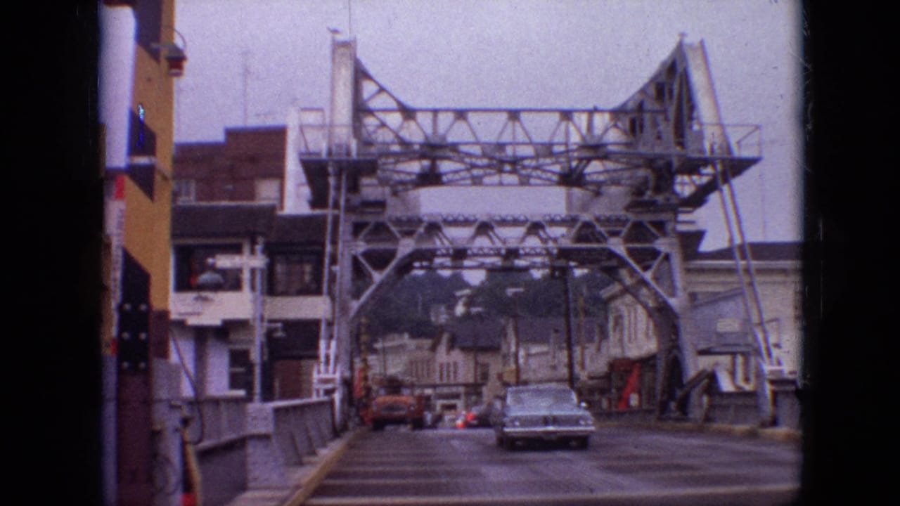 1968 Branson, Missouri 8mm frame showing a light blue vintage sedan driving toward the camera across a steel truss bridge with buildings and distant cars behind it.