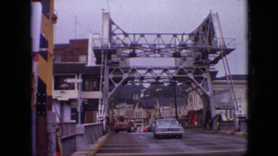 1968 Branson, Missouri 8mm frame showing a light blue vintage sedan driving toward the camera across a steel truss bridge with buildings and distant cars behind it.
