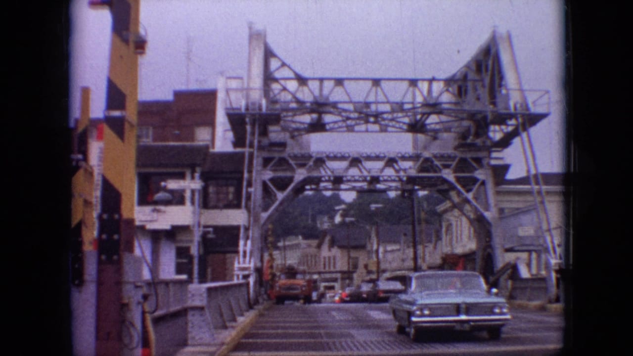 Car nearing the camera on a steel bridge with town buildings in the background.