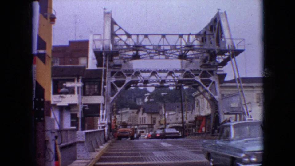 1968 Branson, Missouri home-movie frame showing the steel truss bridge and town street as a vintage car passes near the right side of the frame.