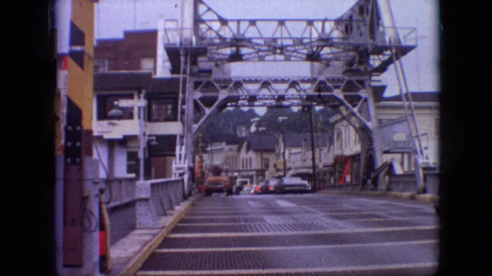 1968 Branson, Missouri frame looking down the steel truss bridge toward town buildings and several distant cars after the nearer vehicle has passed.