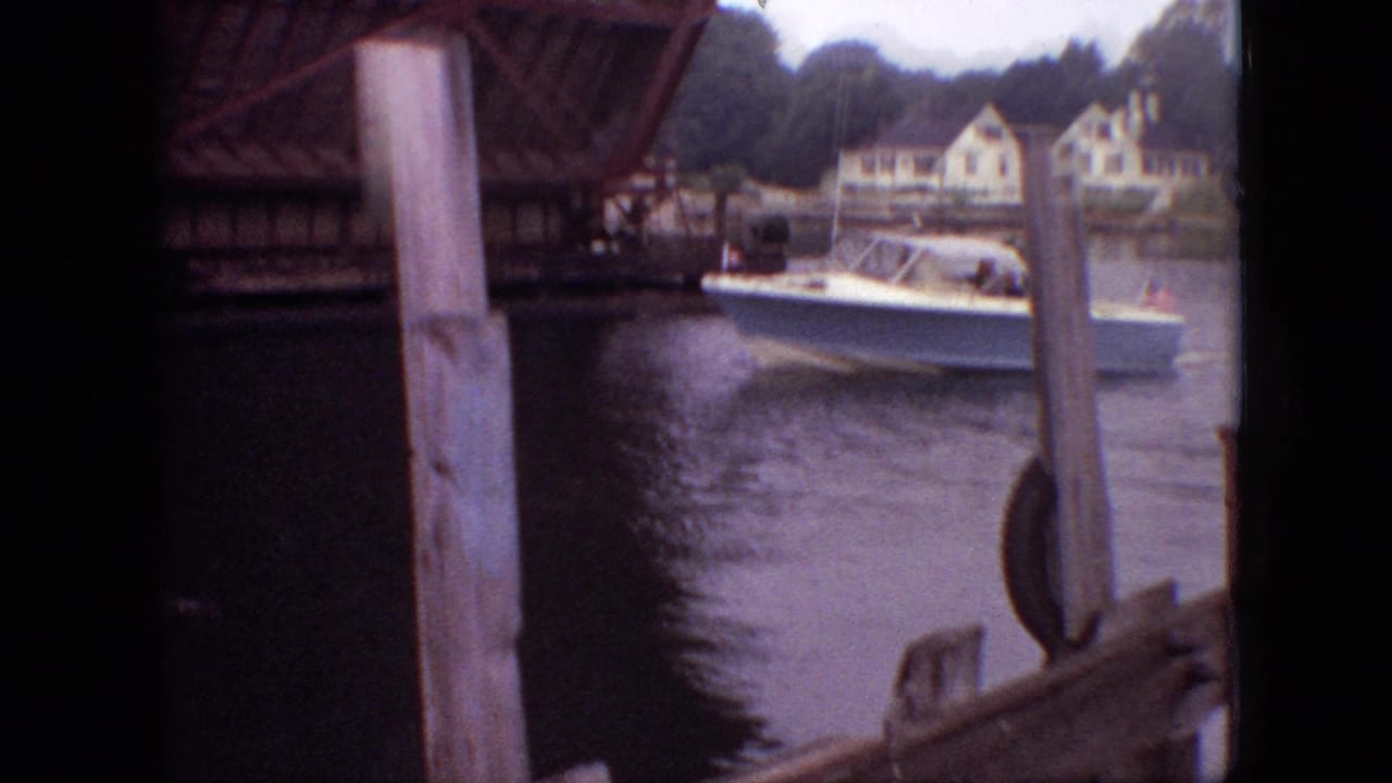 White motorboat on calm water near wooden pilings and a raised bridge.