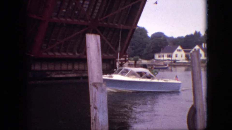1968 Branson, Missouri home-movie frame showing a white motorboat passing through the opened drawbridge area, framed by pilings, bridge steel, water, and houses on the bank.