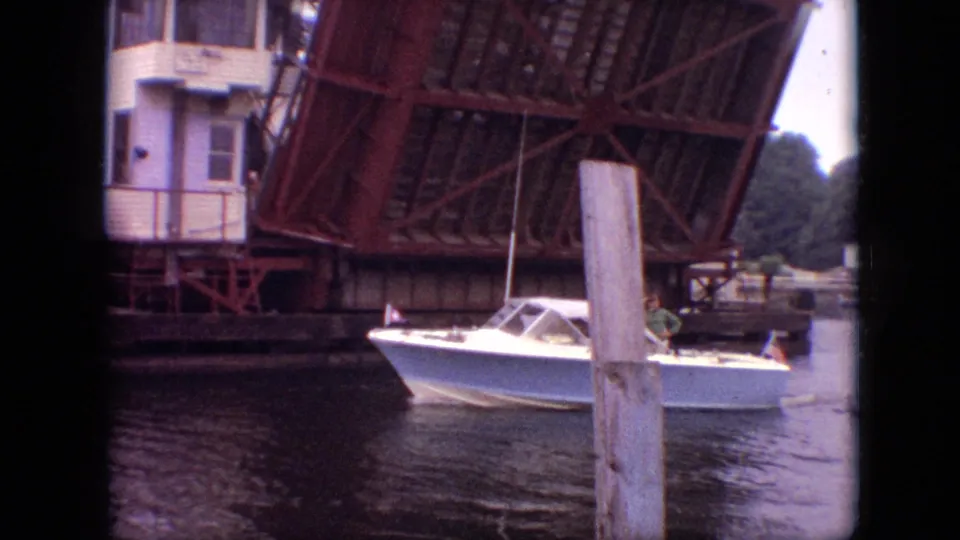 White runabout motorboat continues past the raised red drawbridge in Branson, Missouri, with water, pilings, bridge machinery, and shoreline structures visible.
